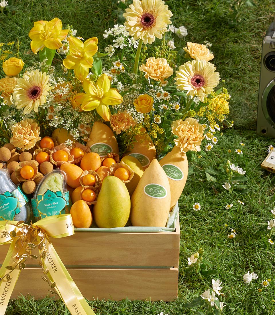 Wooden crate overflowing with tropical Nam Dok Mai mangoes, longans, physalis, and pears with yellow gerbera daisies and
