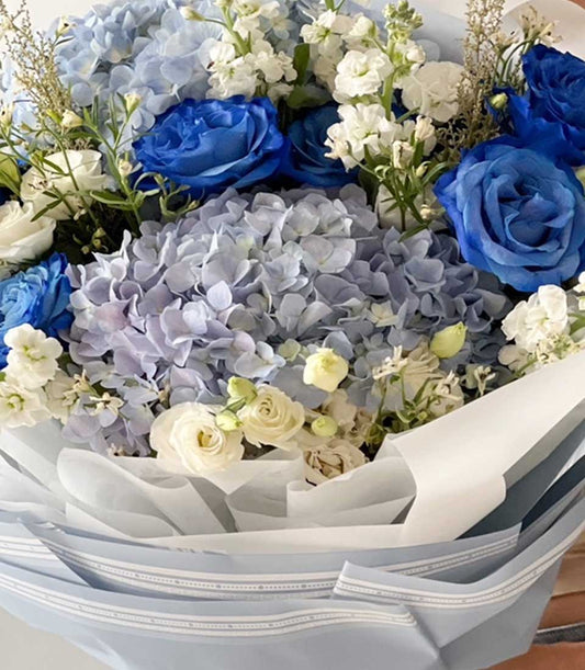 Blue and white fresh flower bouquet with hydrangeas, roses, and white fillers in striped paper wrapping