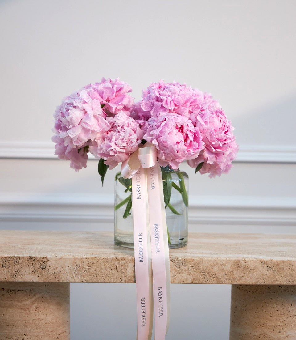 Pink peonies in glass vase with pink ribbon bow on cork shelf against white wall.