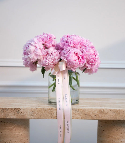 Pink peonies in glass vase with pink ribbon bow on cork shelf against white wall.