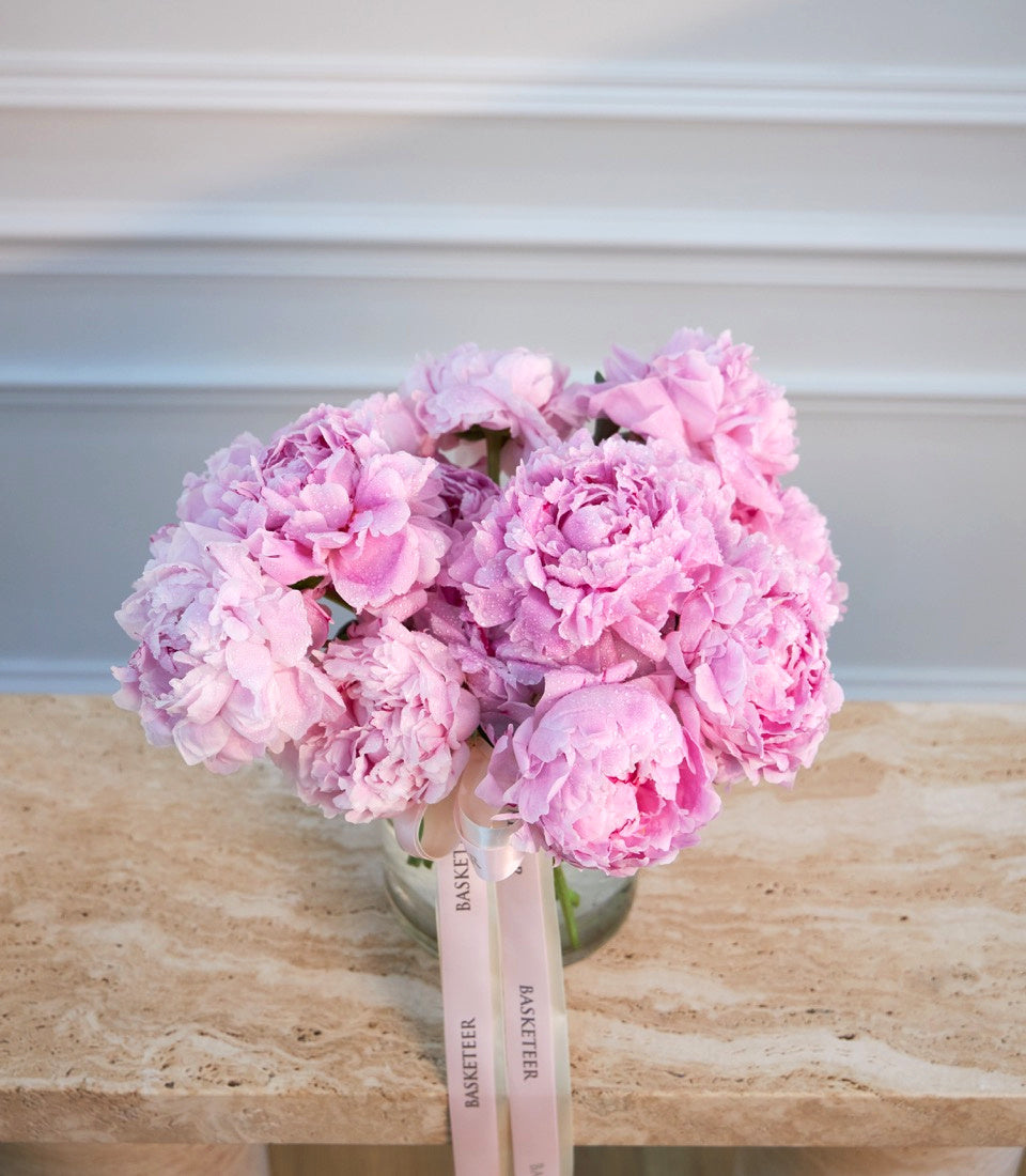 Pink peonies in glass vase with pink ribbon bow on wooden surface against white paneled wall.