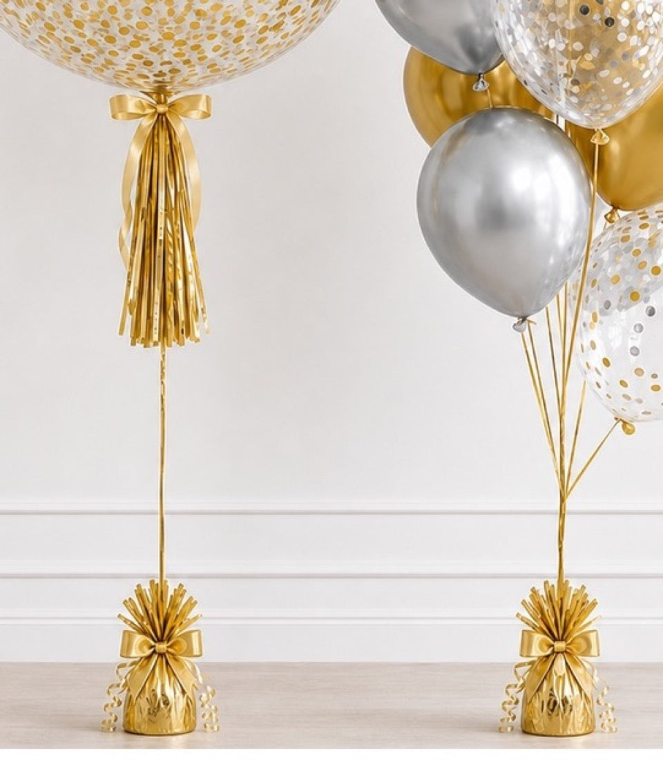 Elegant helium balloons in gold and silver with sequin patterns, gold ribbon bows and tassels, metallic balloon weights on white paneled wall.