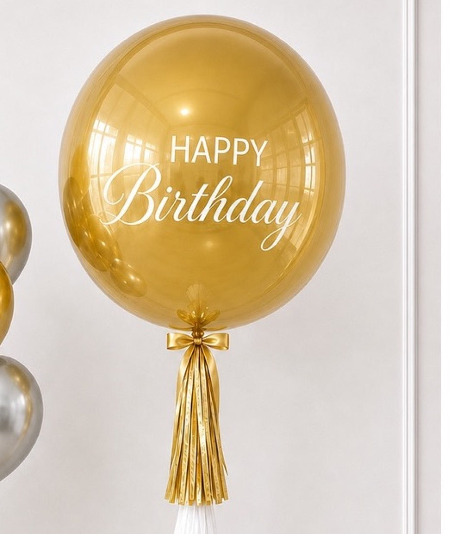 Gold "Happy Birthday" bubble balloon with gold tassel and white streamer tail against white background.