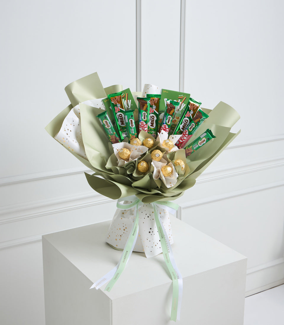 Green and white gift bouquet with snack packets, golden chocolate truffles, and sage wrapping paper in a spotted ceramic vase with ribbon.