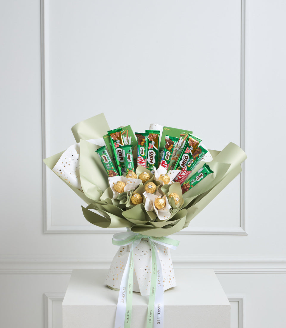 Luxury gift bouquet with green and cream tissue wrapping, golden Ferrero Rocher chocolates, KitKat candy bars, and white ribbon bow on white pedestal.