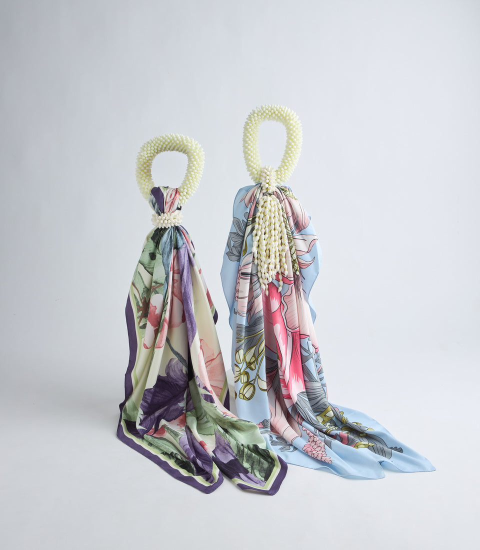 Two decorative silk scarves with floral prints draped from cream-colored beaded loop handles. Left scarf features purple, green, and pink tones; right scarf in blue, pink, and yellow hues.