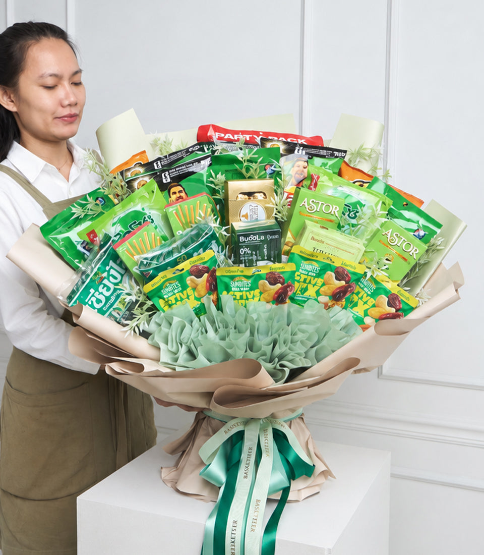 Woman holding large green and cream bouquet featuring snack packages, dried botanicals, and green ribbon bow on white background.
