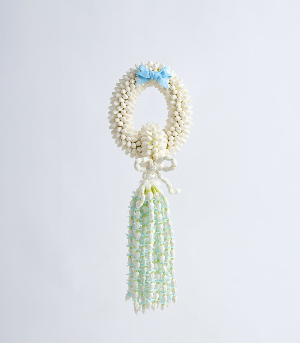 Floral garland necklace with cream daisy wreath, blue bow, and pale green tassel pendant against white background.