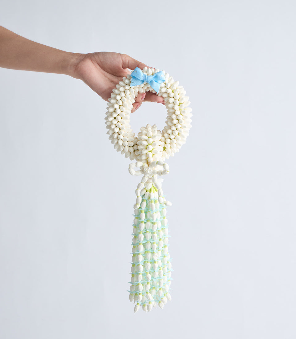 Hand holding white flower garland lei with blue bow and cascading pale green and white flower tail strand.