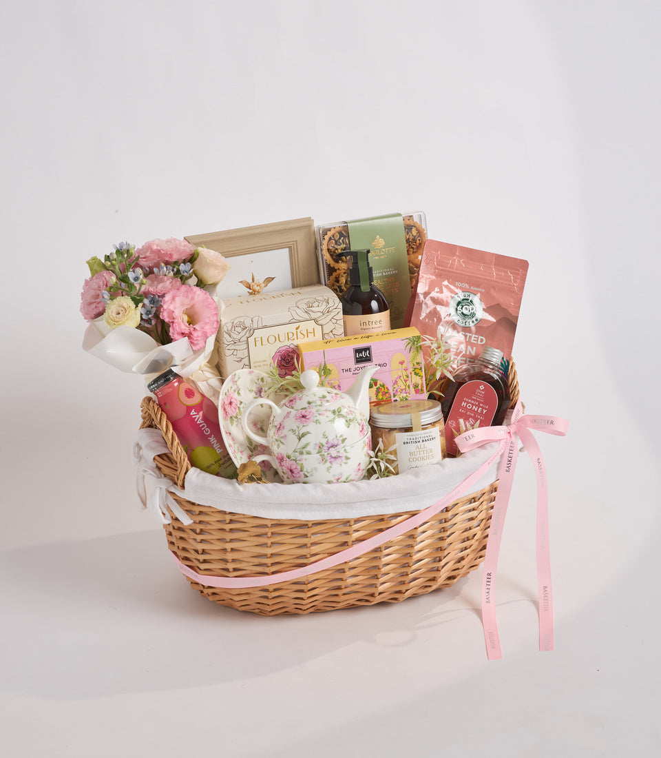 Luxury wicker gift basket with pink flowers, floral teapot, gourmet treats, wine, and decorative items tied with pink ribbons.