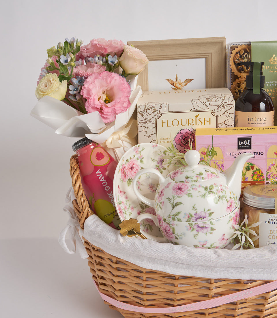 Luxury gift basket with pink floral teapot, fresh flowers, tea, wine, and decorative items in white wicker basket.