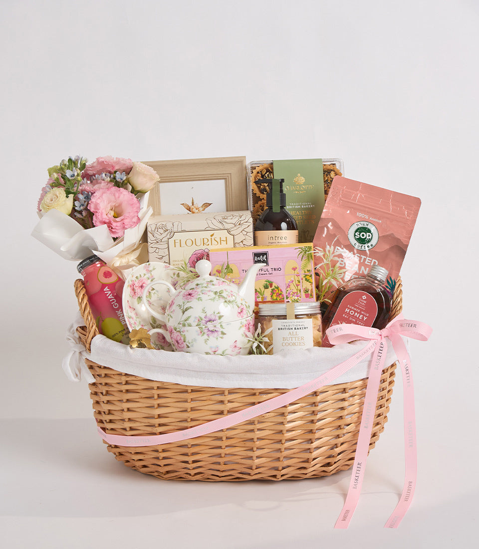 Luxury wicker gift basket with pink flowers, floral teapot, gourmet treats, wine bottle, cookies, and honey jar, tied with pink ribbon.