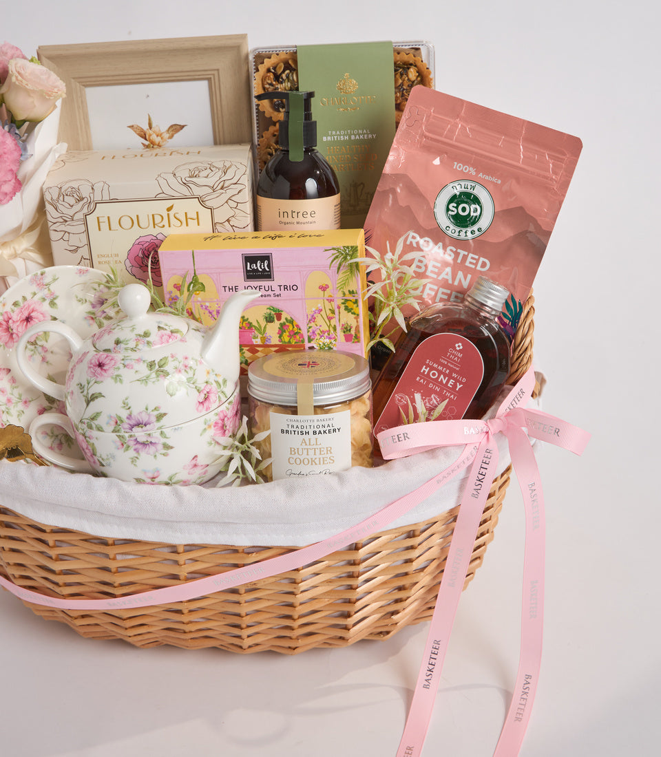Luxury gift basket with wicker weave, pink ribbon, floral teapot, coffee, honey, bakery items, wooden frame, and flowers on white background.