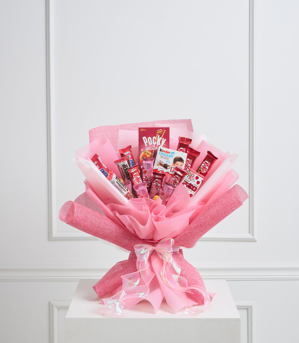 Pink candy bouquet featuring Pocky, Kinder chocolate, and red sweets wrapped in pink tissue paper with pink bow on white pedestal.
