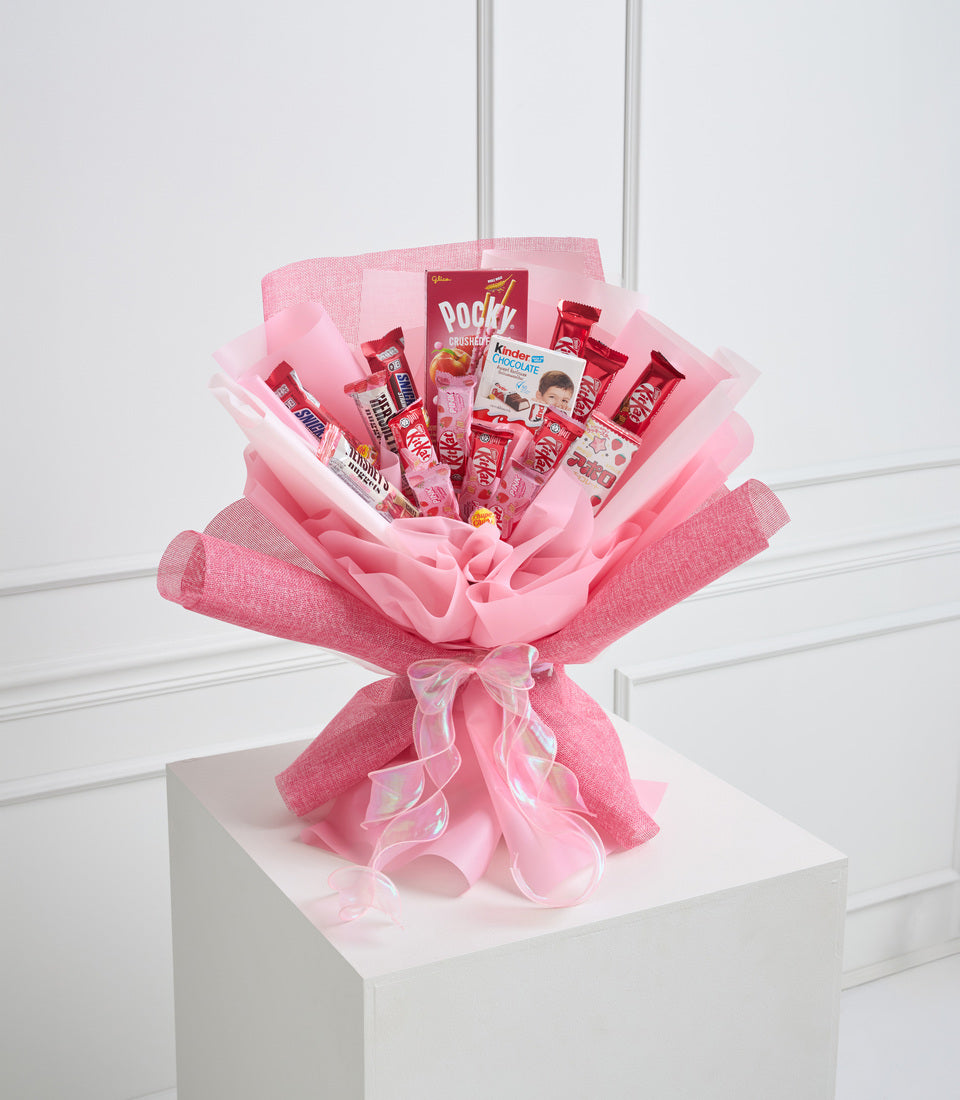 Pink candy bouquet with assorted chocolates and sweets, wrapped in textured pink and light pink paper with ribbon bow, displayed on white cubic stand.
