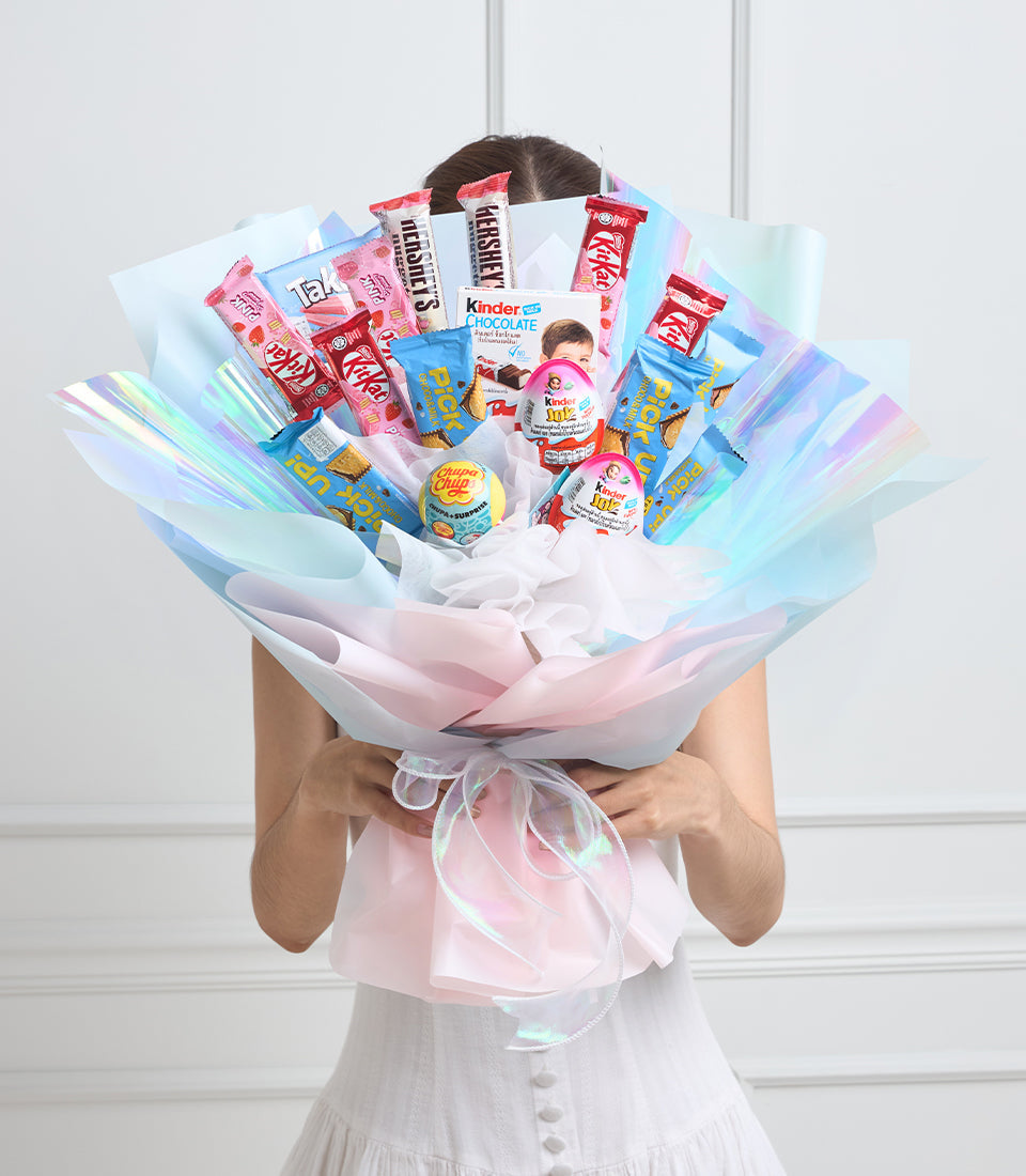 Large candy bouquet with pastel pink, blue and white cellophane wrapping, filled with chocolate bars, Kinder products and lollipops, held by person in white dress.