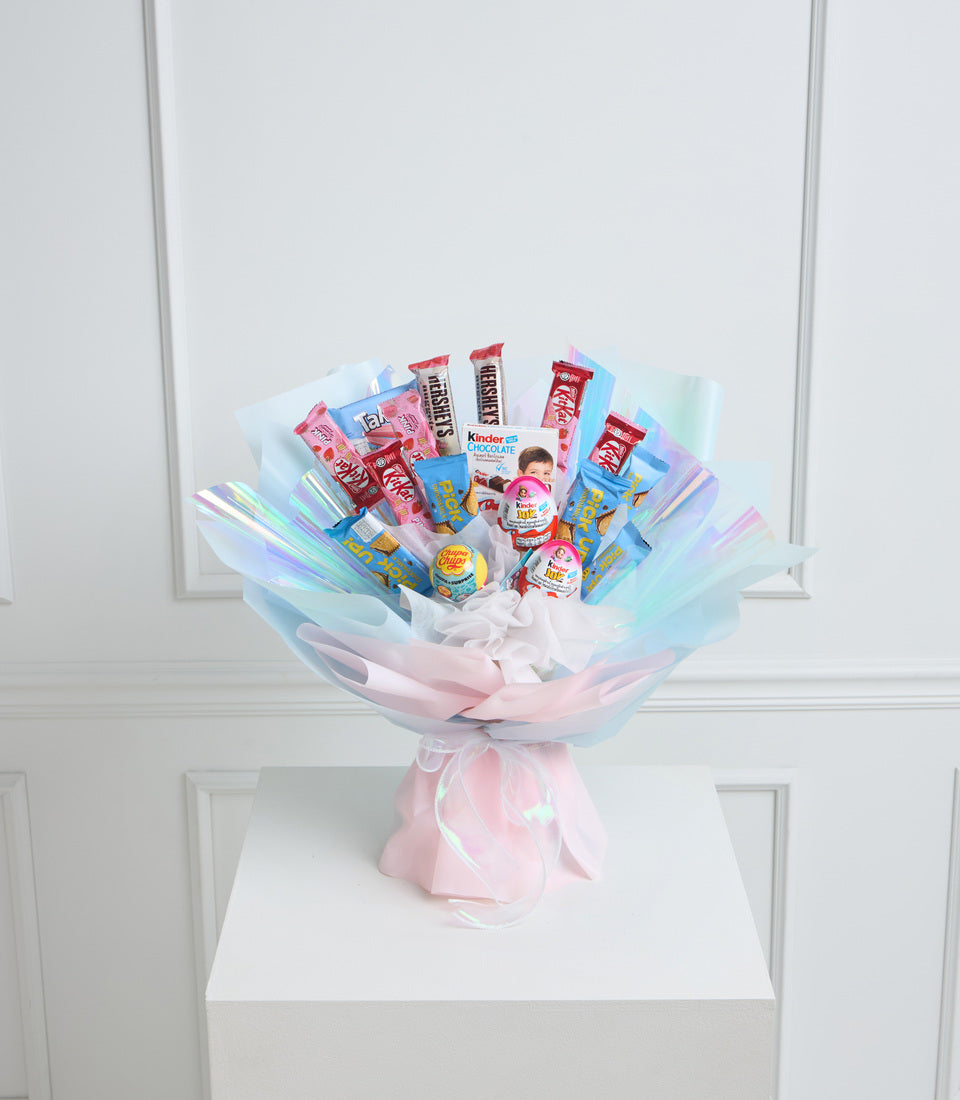 Colorful candy bouquet with pastel pink and blue cellophane wrapping, featuring Hershey's, Kinder, and Lollipop treats arranged in a fan display on white cube pedestal.