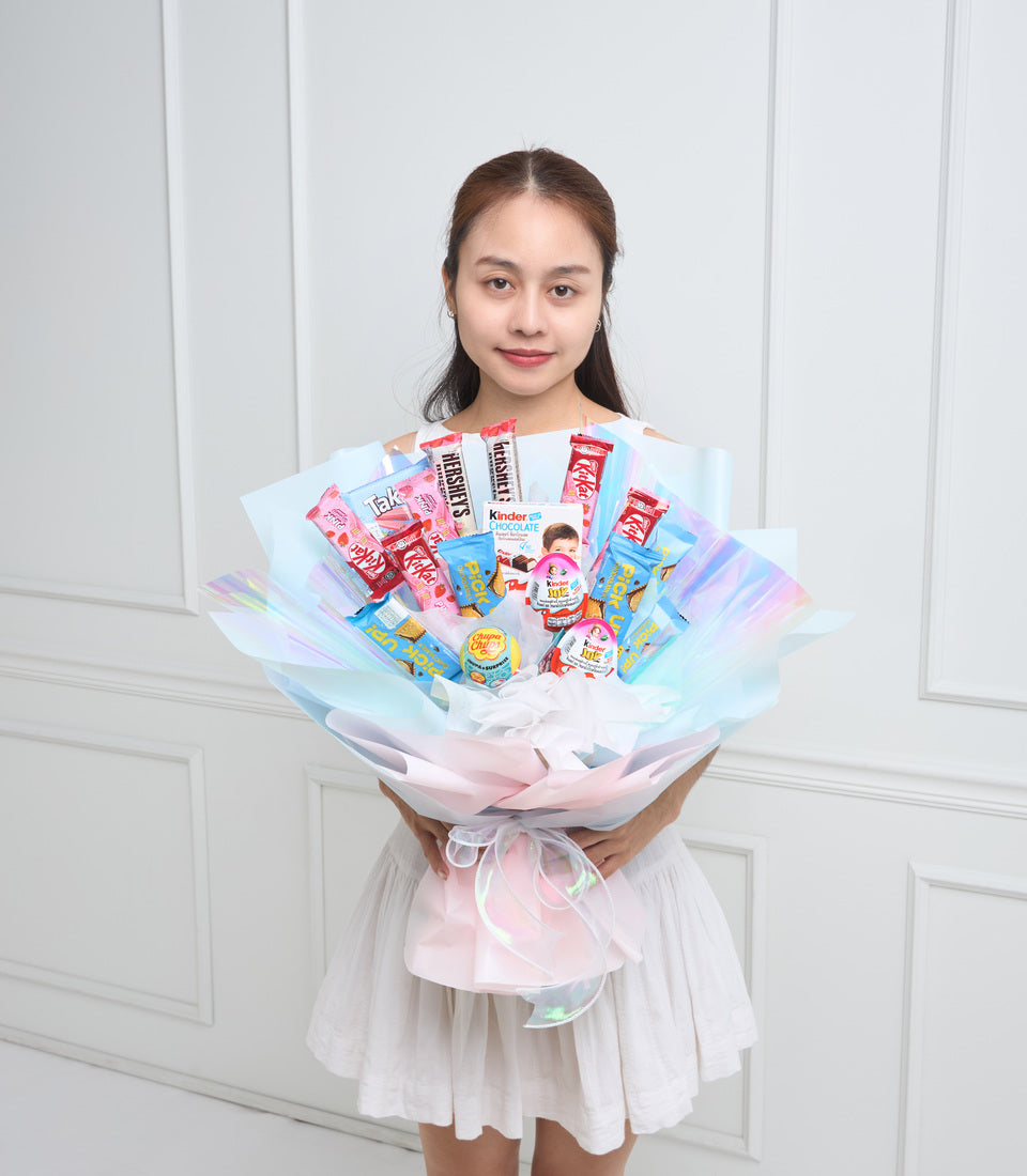 Girl holding colorful candy bouquet with pink and blue cellophane wrapping, featuring chocolate bars and confectionery arranged in fan shape.
