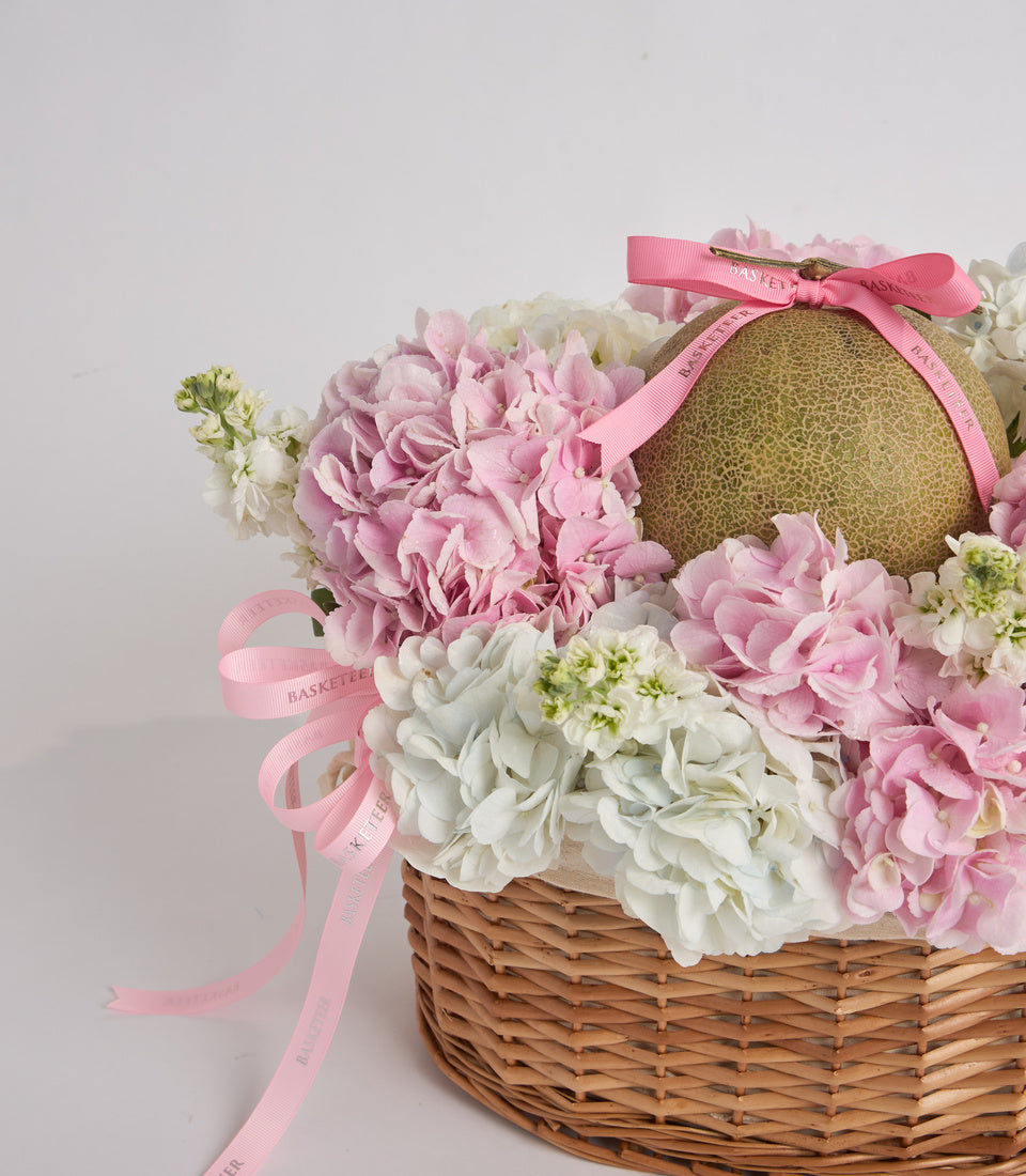 Wicker basket filled with pink and white peonies and hydrangeas, topped with a gold leopard-print box tied with pink ribbon bow.