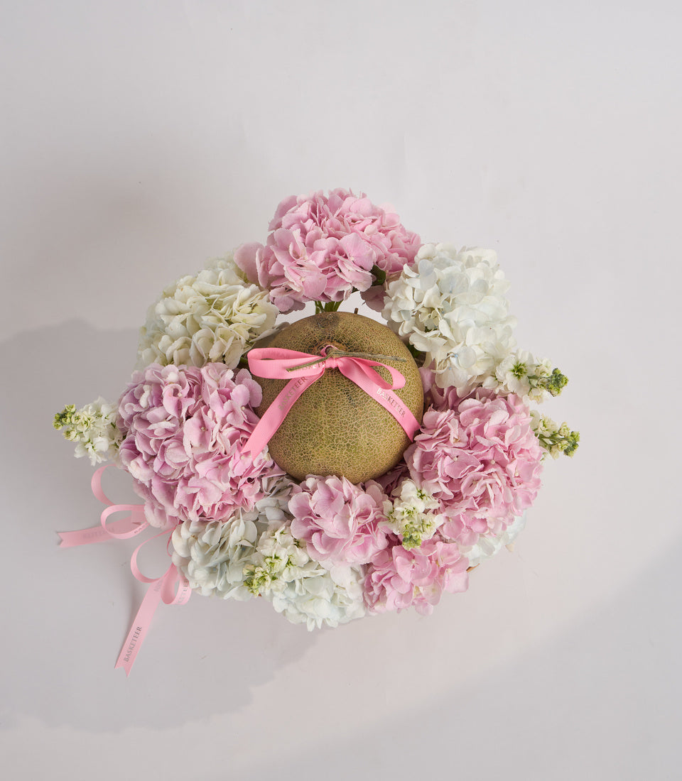Luxurious floral arrangement featuring pink and white hydrangeas with pale greenery, centered around a textured gold sphere adorned with pink ribbon bow.