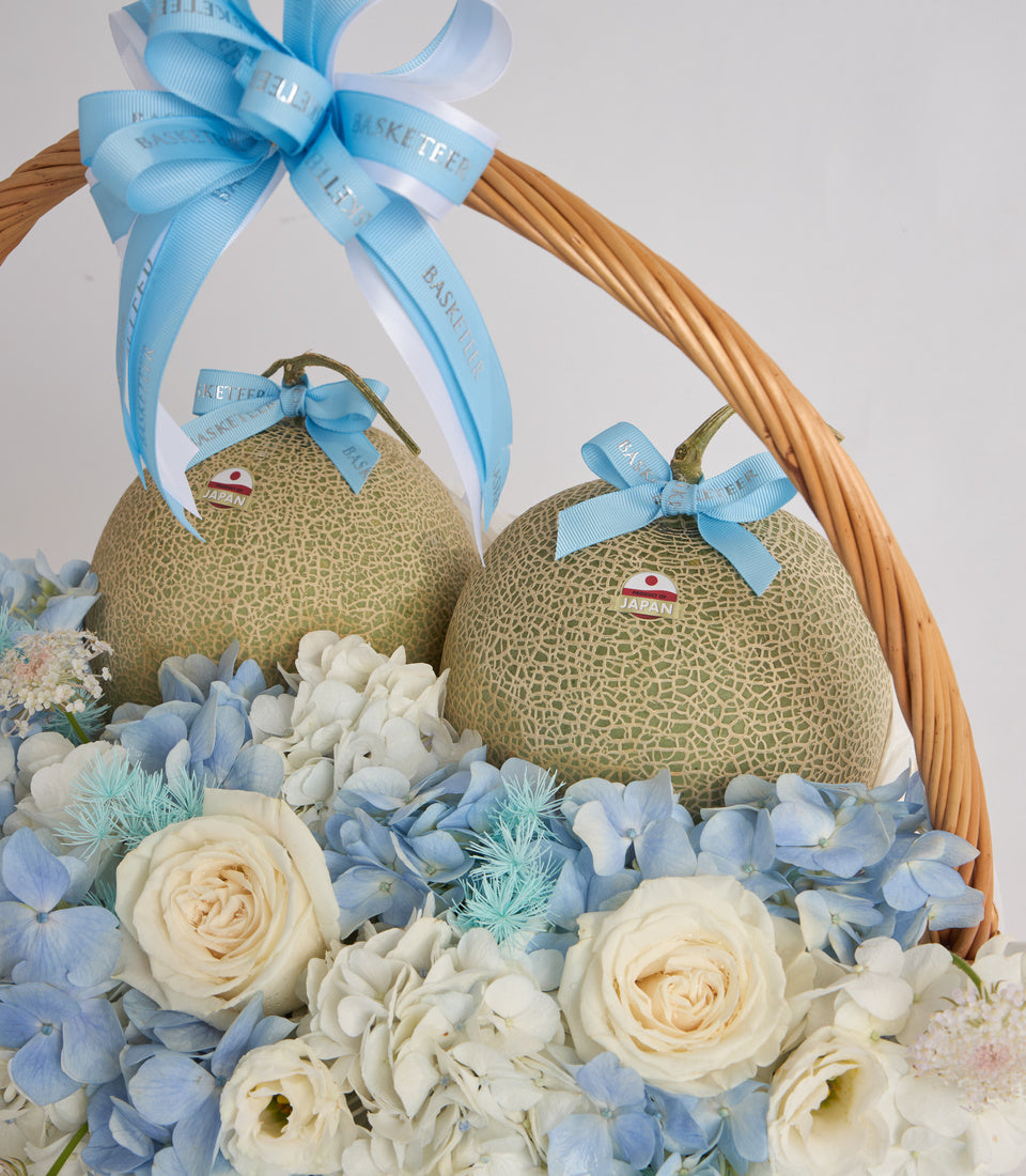 Luxury gift basket with two Japanese melons, blue ribbon bows, cream roses, blue hydrangeas, white flowers, and woven wicker handle.