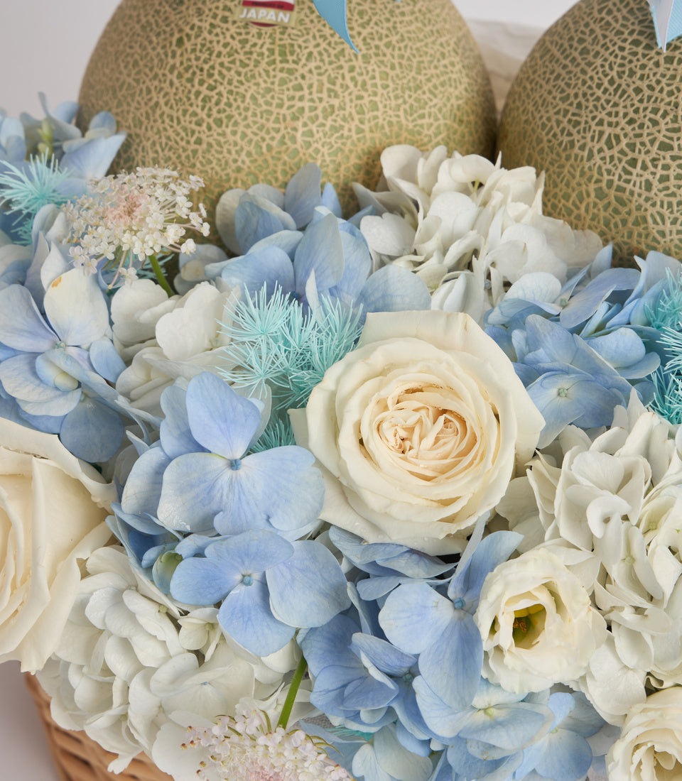 Elegant floral arrangement with white roses, blue hydrangeas, and thistle in woven basket with two Japanese melons behind.