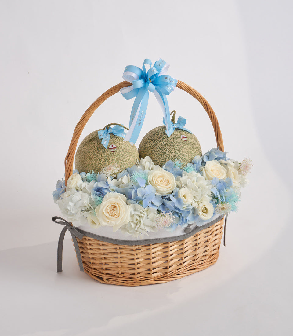# Alt Text

Wicker gift basket with cream roses, blue hydrangeas, white flowers, two melons with blue ribbons, and a blue bow on the handle.