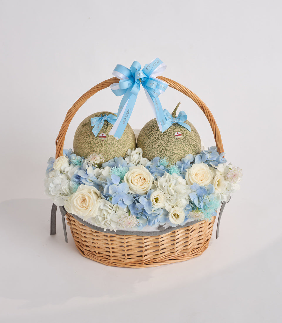 Wicker basket with two melon-shaped fruits, cream roses, blue hydrangeas, white flowers, and blue ribbon bow.