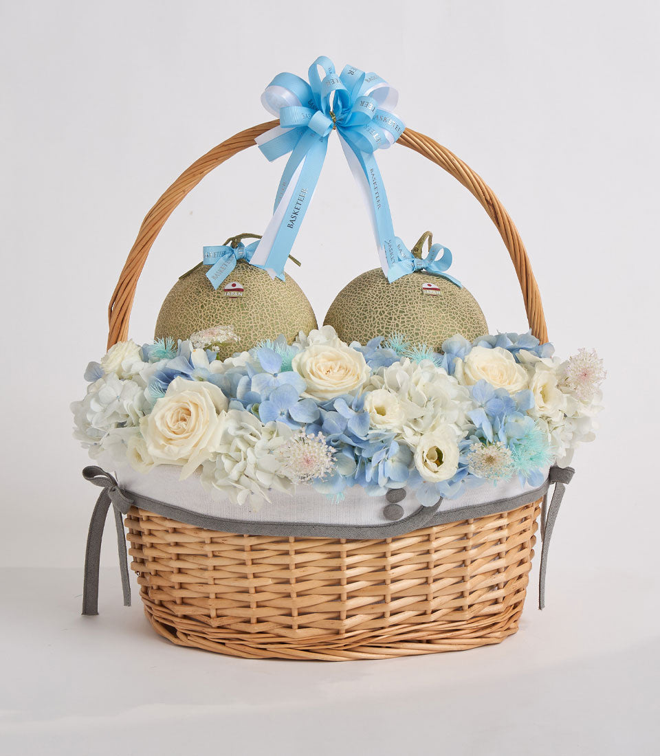 Wicker basket with white flowers, blue hydrangeas, cream roses, two decorative melons with blue bows and gray ribbon handles.