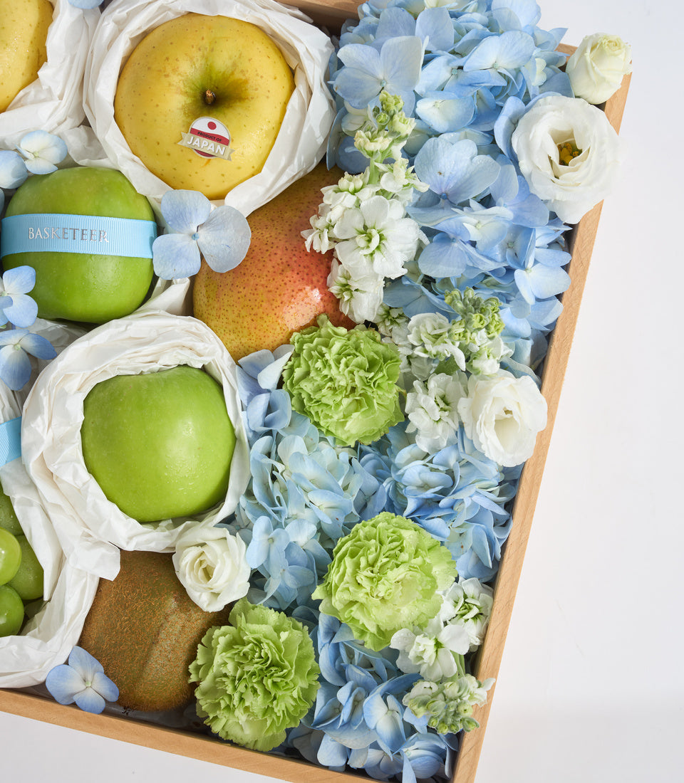 Luxury gift basket with Japanese apples, pears, green carnations, blue and white hydrangeas, and white roses in wooden box.