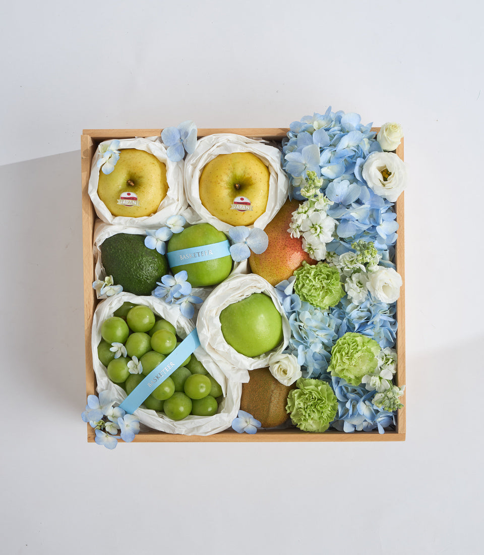 # Alt Text

Wooden gift box with assorted fresh fruits in white paper wrapping, blue and green flowers, featuring apples, grapes, citrus, and green carnations.