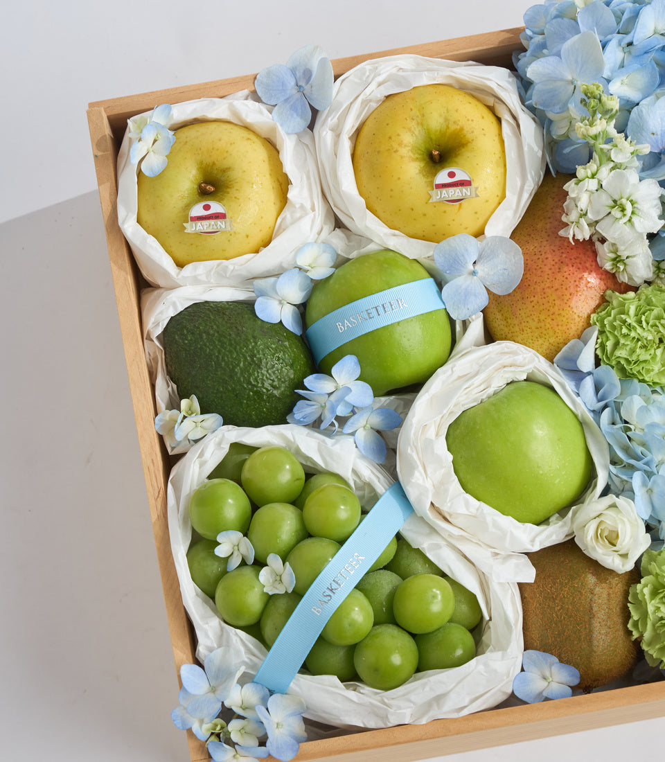 Wooden gift box with premium Japanese fruits: yellow apples, green apples, grapes, and avocado, wrapped in white paper and decorated with blue and white flowers.
