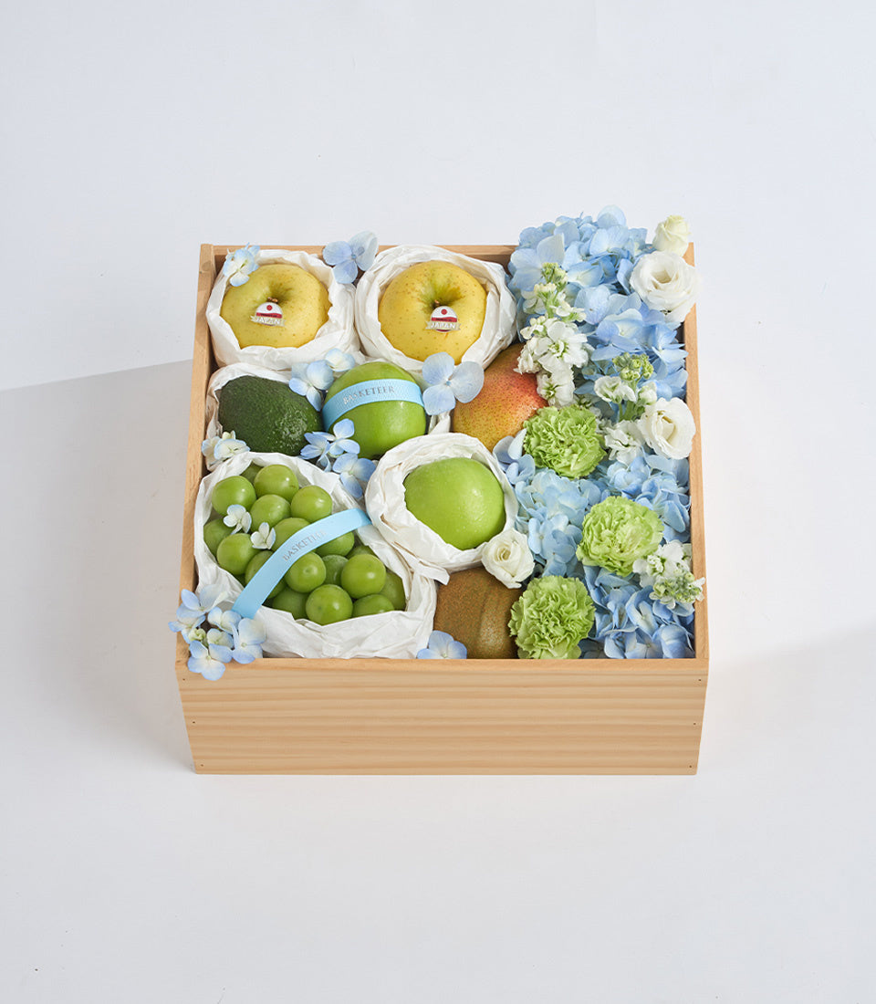 Wooden gift box filled with fresh fruits, green grapes, apples, pears, and decorative blue hydrangeas, white flowers, and cream ribbon accents.