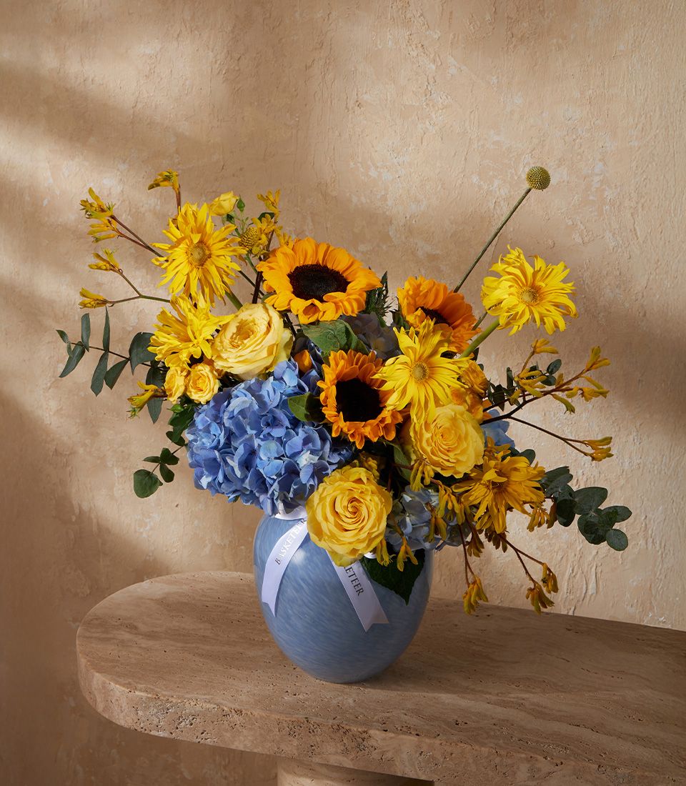 Yellow and blue fresh flower arrangement in light blue glass vase with sunflowers, roses, and hydrangeas