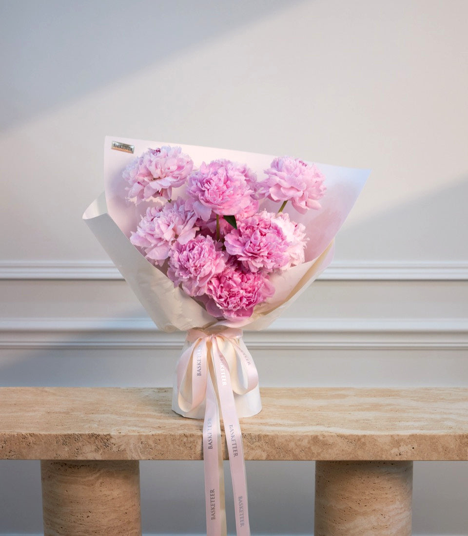 Pink peonies bouquet wrapped in white paper with pale pink ribbon, displayed on a wooden shelf against a white paneled wall.