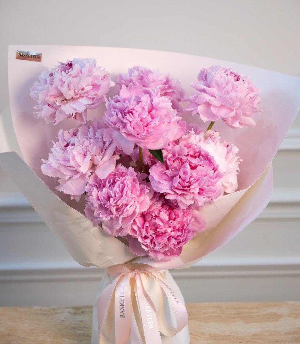 Luxurious bouquet of pink peonies wrapped in soft blush paper with satin ribbons, arranged in elegant floral display.