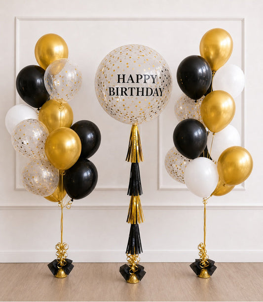 Elegant birthday balloon display with gold, black, and white balloons featuring confetti patterns, centered large "Happy Birthday" balloon with gold and black tassels.