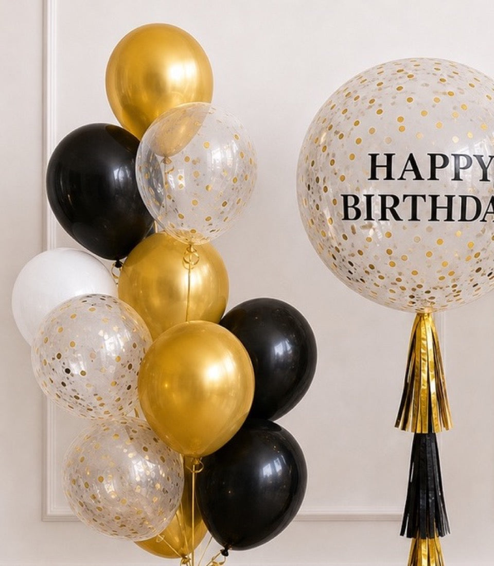 Elegant birthday balloon arrangement featuring clear confetti balloons, gold metallic balloons, black balloons, and gold tassel decorations against a white wall.