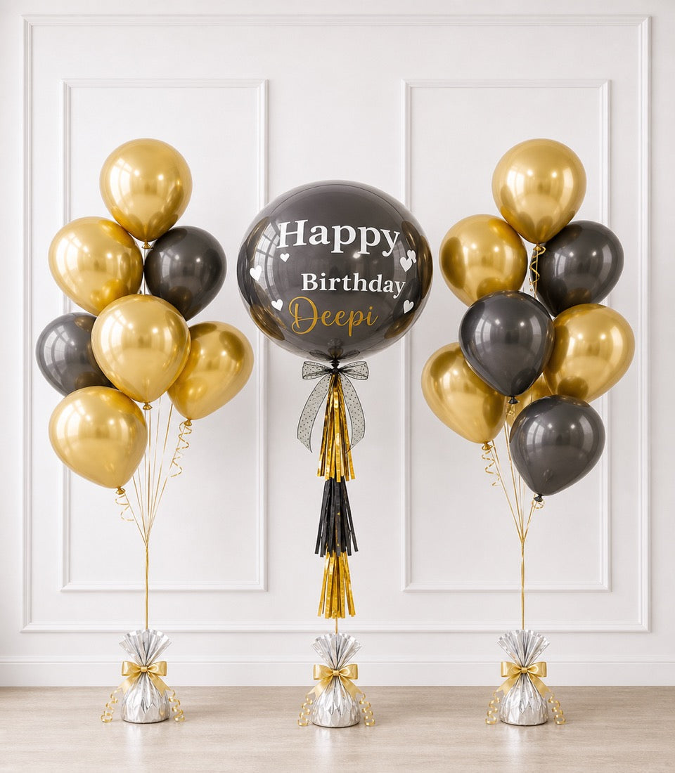 Elegant birthday balloon display with gold and black metallic balloons, central "Happy Birthday Deepi" balloon with tassel, and silver weighted bases with gold bows.