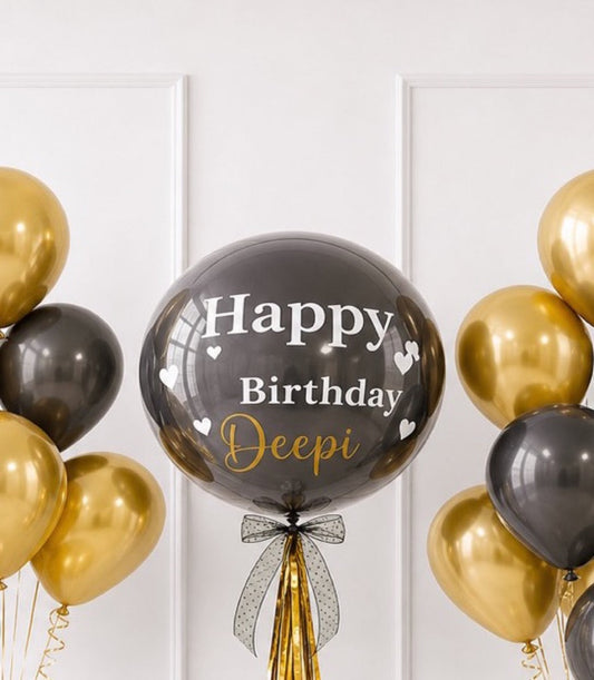Elegant black and gold birthday balloon display with "Happy Birthday Deepi" text, featuring a large central bubble balloon surrounded by metallic gold and black balloons with decorative bow.