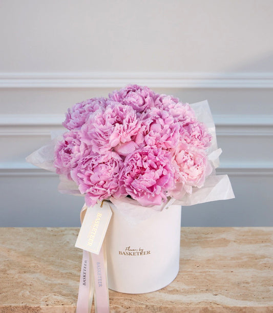 Pink peonies in a white cylindrical vase with tissue paper wrapping and a pale pink ribbon bow, branded "Flowers by Basketeer."