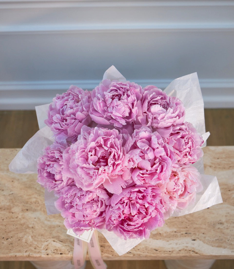 Pink peonies bouquet wrapped in white tissue paper with pink ribbon, displayed on a wooden surface against a light gray wall.