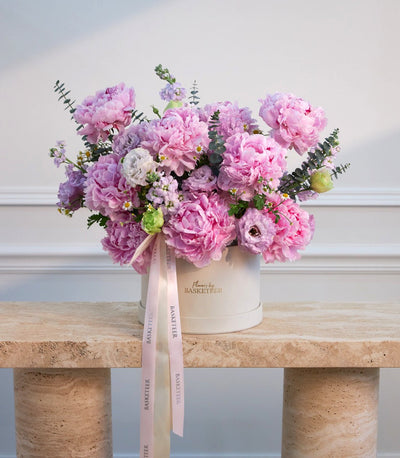Luxurious pink peony bouquet in cream cylindrical box with pale pink ribbon, displayed on natural cork table against white paneled wall.