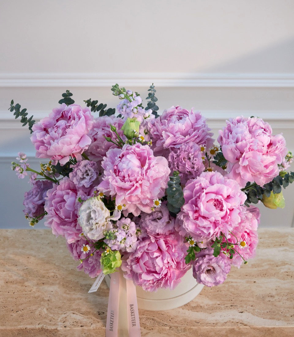 Luxurious bouquet of pink and purple peonies with white blooms, eucalyptus, and chamomile flowers in a cream ceramic vase tied with pink ribbon, displayed on a beach.