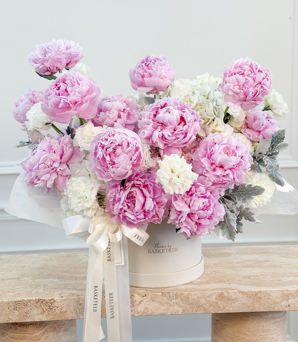 Luxurious pink and white peonies in a cream cylindrical vase with "Basketeer" branding and cream silk ribbons on wooden surface.