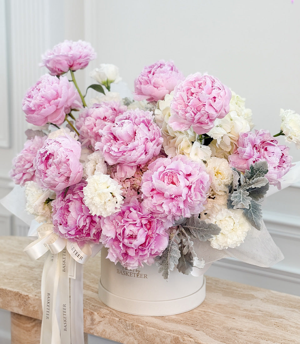 # Alt Text

Luxurious pink and white peonies with grey-green foliage in a white cylindrical vase with cream ribbon bow on wooden surface.