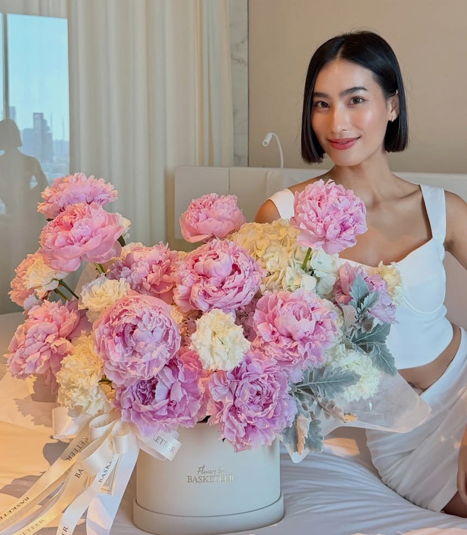 Luxurious pink and cream peonies in white cylindrical vase with gold ribbons, displayed by woman in white tank top.