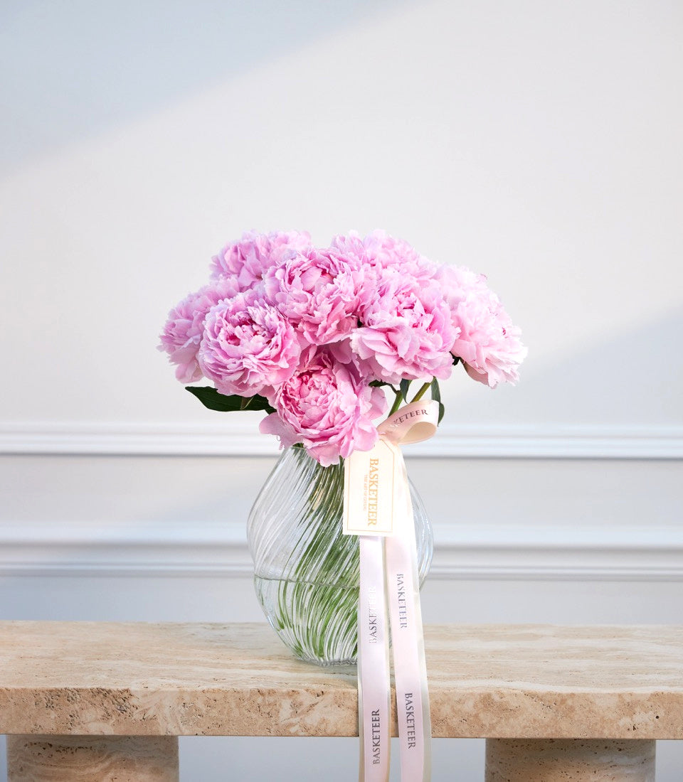 Pink peonies in a clear ribbed glass vase with a pink ribbon bow on a wooden surface against a white paneled wall.