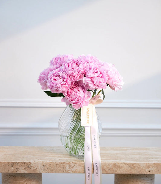 Pink peonies in a clear ribbed glass vase with a pink ribbon bow on a wooden surface against a white paneled wall.