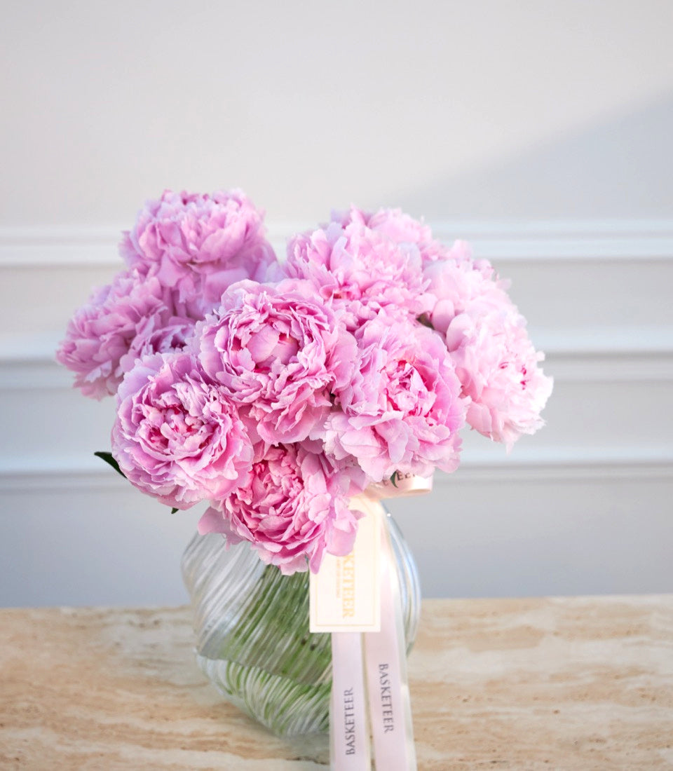Pink peonies in clear glass vase with green stems and white ribbon bow on wooden surface against white wall.